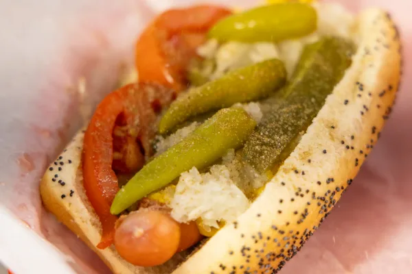 Hot dog topped with pickle spears, tomato, and peppers in a poppy seed bun